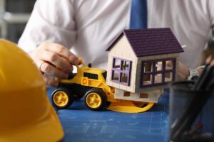 una mano gioca con un modellino di una ruspa e quello di una casa